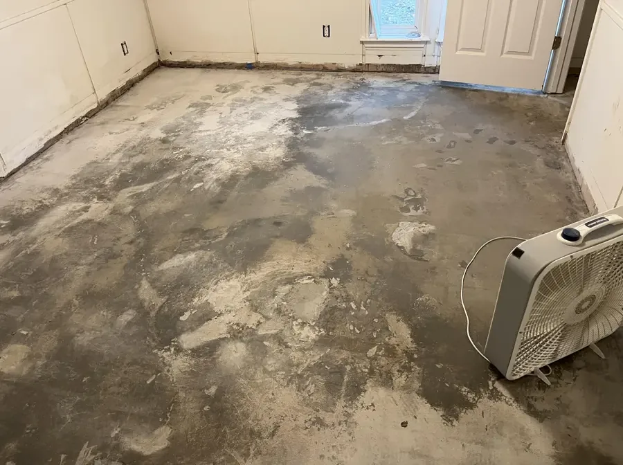 Bare concrete slab with dark wet patches and a fan running after the flooring came up